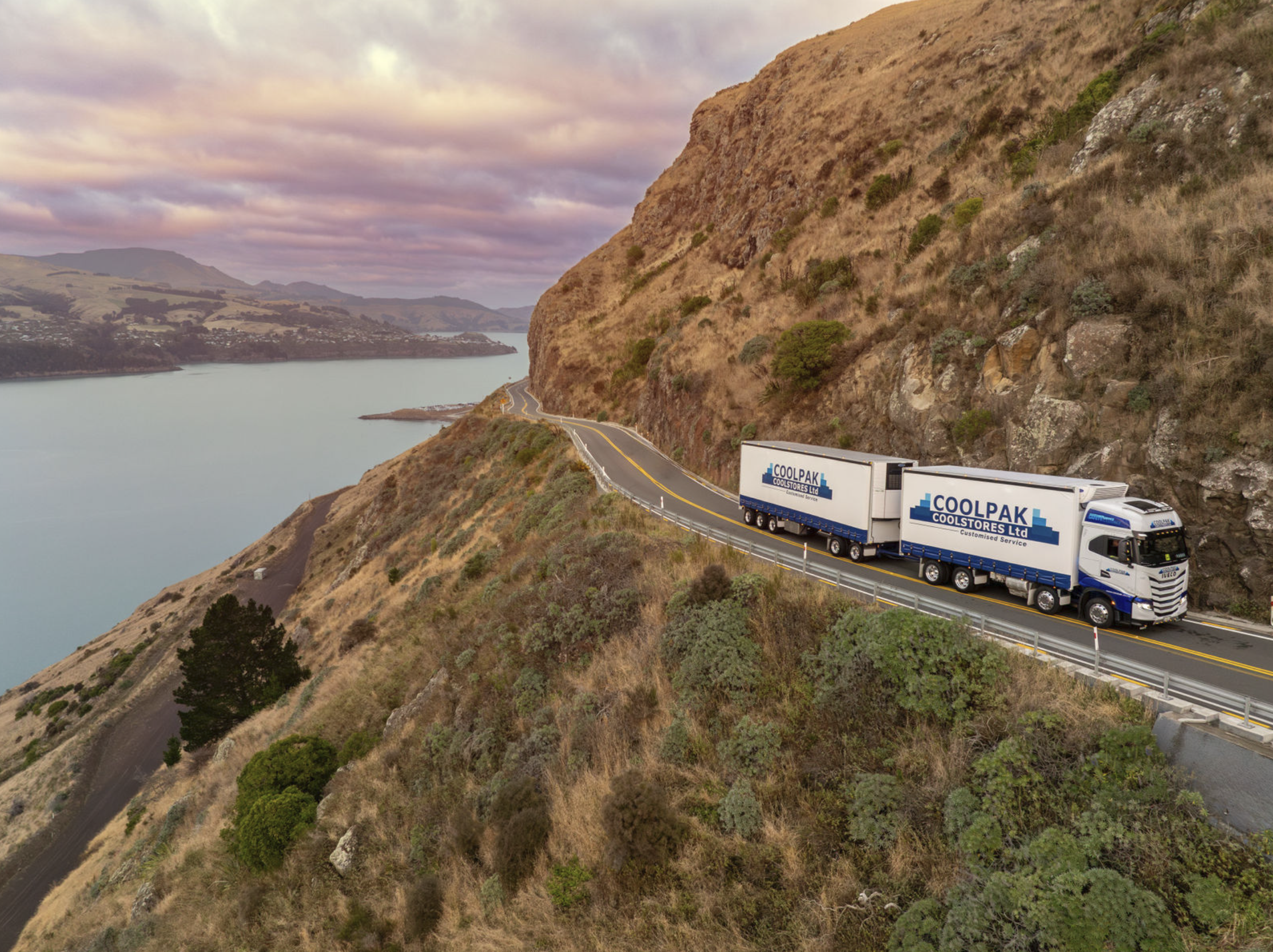 A CoolPak Coolstores semi-truck driving along a winding coastal road surrounded by hills and water