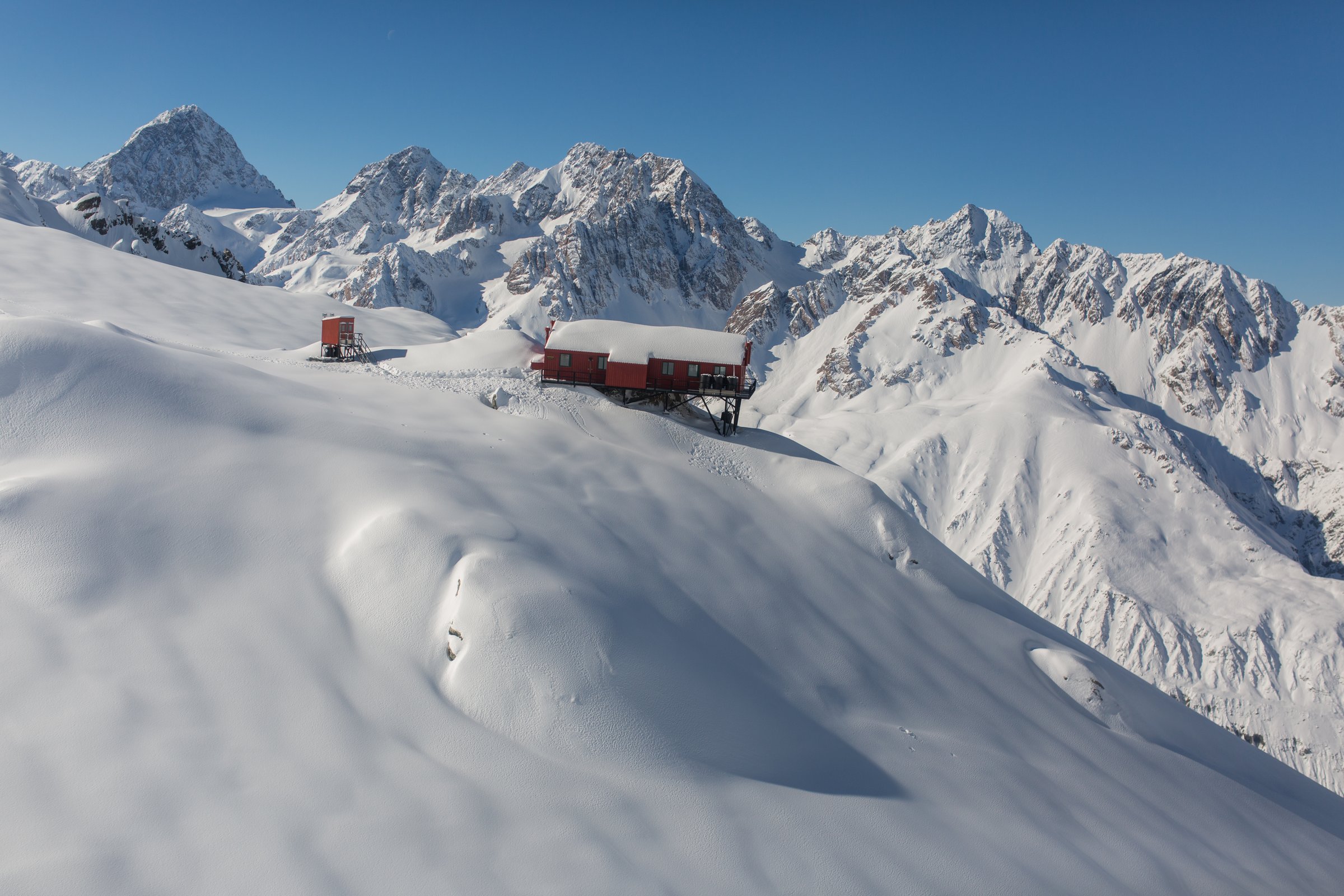 Aerial view of a red alpine hut perched on a deep snow ridge in the Southern Alps