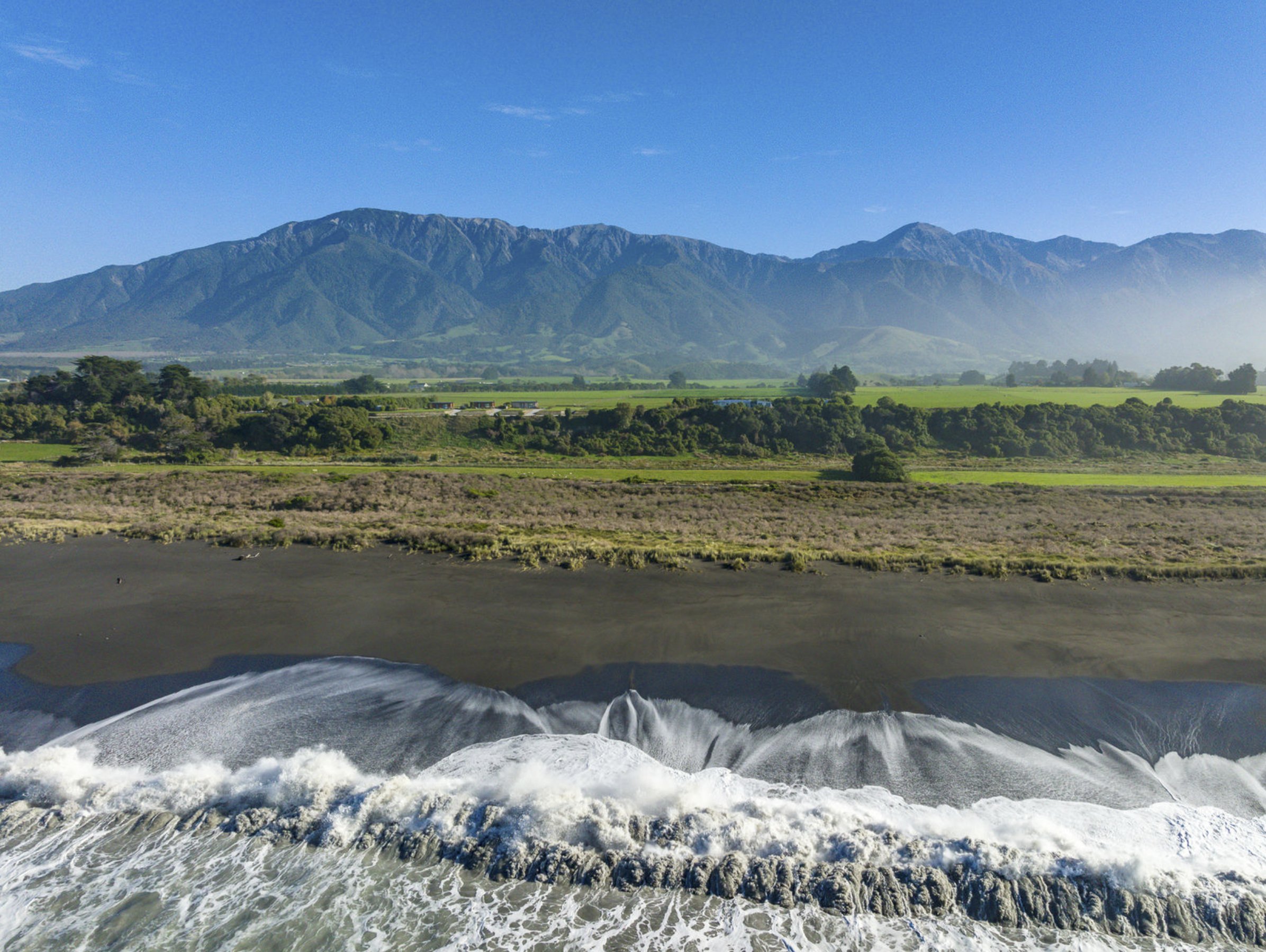 Aerial view of surf breaking on a dark sand beach with farmland and coastal mountains behind