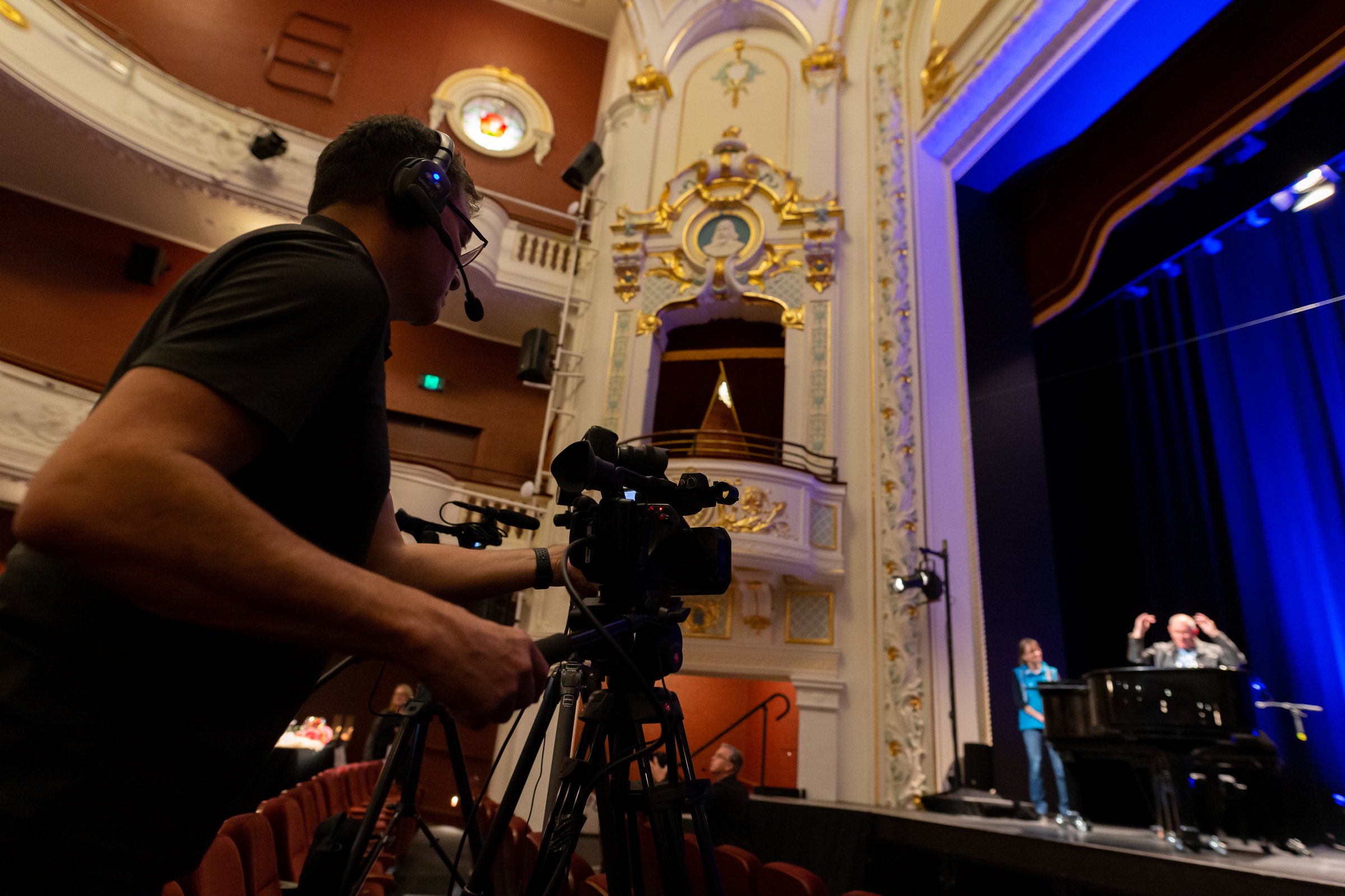 Camera operator filming a rehearsal beneath the ornate balconies of a heritage theatre
